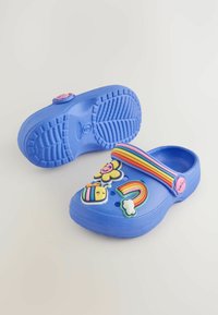 Blaue Clogs mit einer strukturierten Gumm Oberfläche, ausgestattet mit einem Regenbogenriemen und bunten plastikverzierungen, darunter Blumen, eine Biene und Wolken.