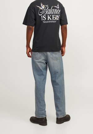 Personne portant un t-shirt noir avec le texte "L'équilibre est la clé" et des papillons, un jean bleu clair et des chaussures foncées, debout dos tourné vers l'avant.