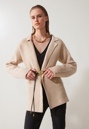 Béžový pletený kardigán so širokými klopami, opásaný čiernym povrázkom a zlatými prvkami. Nosený cez čierny top, v kombinácii s tmavými džínsami.