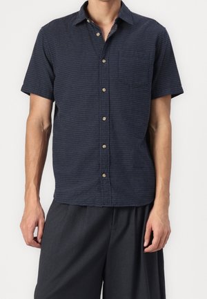 Homme portant une chemise boutonnée à manches courtes, bleu foncé texturée avec une poche poitrine, et un pantalon ample gris foncé, sur un fond uni.