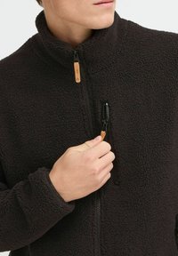 Veste en polaire noire avec un col montant, fermeture éclair à l'avant et poche poitrine. Elle présente un tissu texturé et un accent en bois sur la tirette de la fermeture éclair.