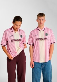 Lyserøde, stribede polo shirts med en cremefarvet midte og en sort krave, med logo og teksten "SWARM" i guld. Modellerne bærer afslappede bukser.