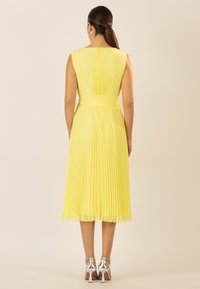 Gelbes plissiertes Kleid mit einem taillierten Bund, knielang, ärmelloses Design und einem verdeckten Reißverschluss auf der Rückseite. Silberne Absätze vervollständigen den Look.