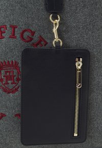 Porte-cartes en cuir noir avec fermeture éclair dorée fixé à un tissu gris avec des lettres rouges brodées et un blason.