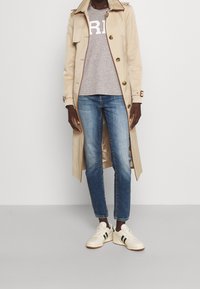 Beige trench coat med bruna accenter, grå T-shirt med vit tryck, blå denimjeans och vita sneakers med gröna ränder och en strukturerad sula.