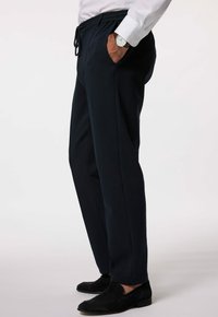 Mężczyzna w czarnych dopasowanych spodniach dresowych, czarnych loafersach, białej koszuli wizytowej i srebrnym zegarku na nadgarstku, stojący z jedną ręką w kieszeni.