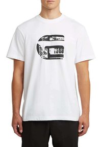 Hombre vestido con camiseta blanca de manga corta con gráfico abstracto negro y texto parcial, combinada con pantalones negros, de pie contra un fondo blanco.