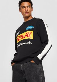 Svart sweatshirt med en iögonfallande gul och röd "REPLAY" logotyp, vita sidokanter och en strukturerad finish. Bärs med mörk denim.