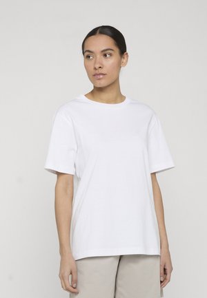 Kvinde med mørkt hår iført hvid kortærmet T-shirt og beige bukser, der kigger til venstre mod en ensartet lys baggrund.