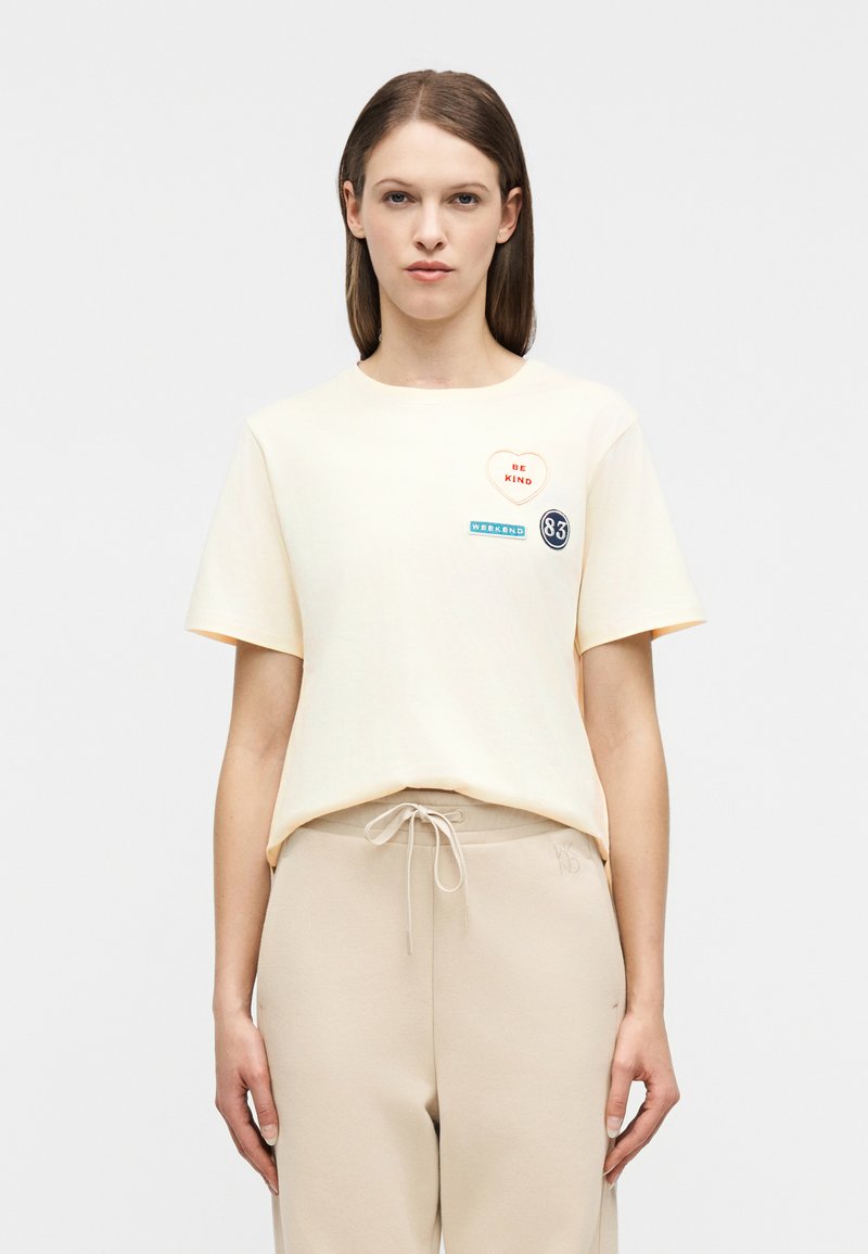 Γυναίκα που φοράει μια κρεμ κοντομ άνικη μπλούζα με μπαλώματα "ΝΑ ΕΙΣΑΙ ΕΥΓΕΝΙΚΗ", "ΣΑΒΒΑΤΟΚΥΡΙΑΚΟ" και "83", συνδυασμένη με μπεζ παντελόνια με κορδόνι στη μέση.