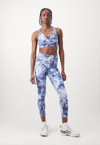 Mėlynas raštas „tie-dye“ sportinis liemenukas ir leginsai, pagaminti iš lygaus audinio. Turi žemą iškirptę ir subtilią ženklinimą kairėje krūtinės pusėje.
