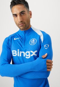 Niebieska longsleeve koszulka sportowa z zamkiem przy kołnierzu, z teksturowanymi liniami i białym logo Nike. "Bingx" wyraźnie nadrukowane na białym tle.