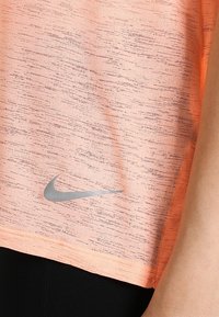 Top de entrenamiento Nike en coral con un acabado texturizado, con un logo plateado en la parte inferior izquierda. Mangas cortas y ajuste relajado.