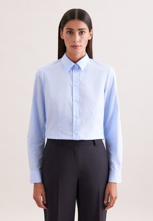 Vrouw met lang donker haar draagt een lichtblauw overhemd met knopen, ingestopt in een zwarte broek met hoge taille, staand tegen een effen achtergrond.