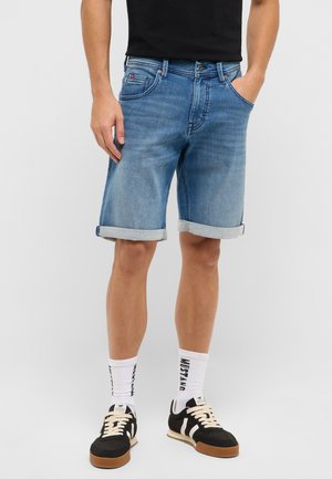 Mann mit hochgekrempelten blauen Jeansshorts, weißen Socken mit schwarzem Text und schwarz-weißen Sneakern vor einem einfarbigen Hintergrund.