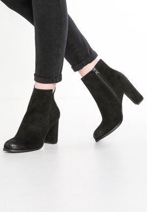 Schwarze Wildleder-Stiefeletten mit Blockabsätzen und seitlichen Reißverschlüssen, getragen zu hochgekrempelten schwarzen Skinny-Jeans vor weißem Hintergrund.