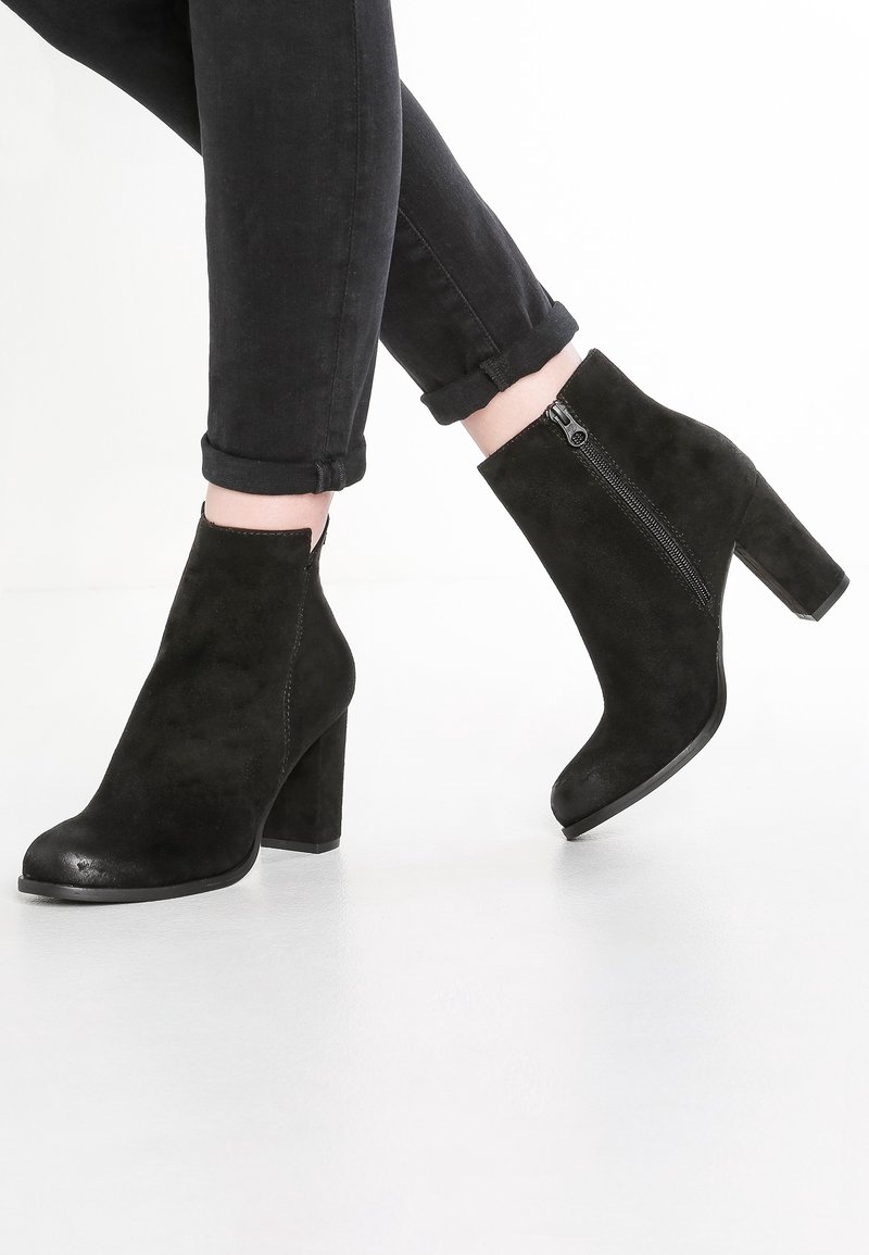 Bottines noires en daim avec talons carrés et fermetures éclair latérales, portées avec un jean skinny noir retroussé, sur fond blanc.