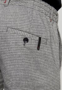 Pantalon à motif pied-de-poule gris avec une texture lisse, doté d'une poche latérale, d'un bouton noir et d'une petite étiquette rouge en accent.