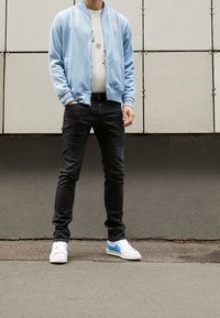 Giacca azzurro chiaro con zip, t-shirt grigia con grafica, jeans neri e sneakers bianche con dettagli blu. Tessuto liscio e vestibilità comoda.