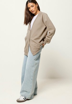 Kobieta w beżowym, oversize kardiganie na guziki, białym t-shircie, szerokich jasnoniebieskich jeansach i szarych sneakersach, stojąca z rękami w kieszeniach.