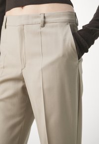 Gros plan sur un pantalon beige sur mesure avec passants de ceinture, pli avant, et une main dans la poche droite, porté avec un haut à manches longues foncé.