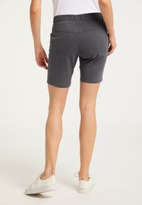 Persona che indossa pantaloni corti grigio scuro fino al ginocchio e sneakers bianche, in piedi contro uno sfondo chiaro e uniforme.