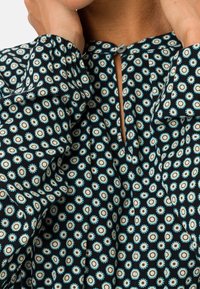 Černá látková blůza se květinovým vzorem v tyrkysové a bílé barvě, s detaily klíčové dírky na krku a nařasenými rukávy.