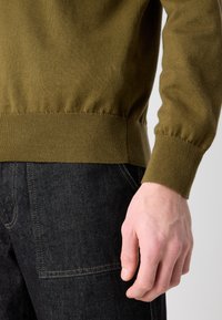 Olive zielenony sweter dzianinowy z ściągaczami przy mankietach i dolnej krawędzi, zestawiony z ciemnymi dżinsami z widocznymi szwami i kieszenią.