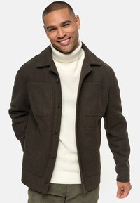 Chaqueta de lana marrón con cuello, dos bolsillos frontales y cierre de botones. Llevada sobre un suéter de cuello alto color crema.