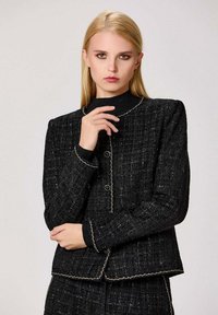 Veste en tweed noire avec un tissu texturé, col rond et coutures contrastées. Fermeture à boutons et manches longues.