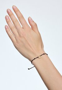 Czarny sznurkowy bransoletka z złotym okrągłym akcentem, zabezpieczona regulowanymi węzłami, noszona na ręce w neutralnym odcieniu skóry na jasnym tle.