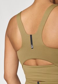 Oliven grønn sportbralette med utskjært bakdesign. Laget av stretchy stoff, med en svart logotag på midten av ryggen.