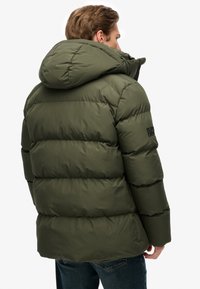 Donkergroene pufferjas met een capuchon en een gequilt patroon. Voorzien van een ritssluiting en zijzakken, gemaakt van een gladde, gestructureerde stof.