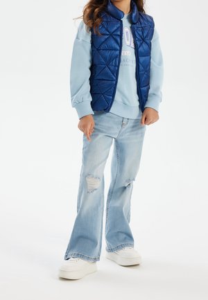 Sininen toppaliivi timanttikuviolla vaaleansinisen collegepaidan päällä, yhdistetty vaaleansinisiin revittyihin liehukkeisiin farkkuihin ja valkoisiin tennareihin.