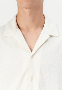 Hvid kortærmet skjorte med et tekstureret mønster, spidse krave og knaplukning. Stoffet har et blødt udseende og en subtil glans.