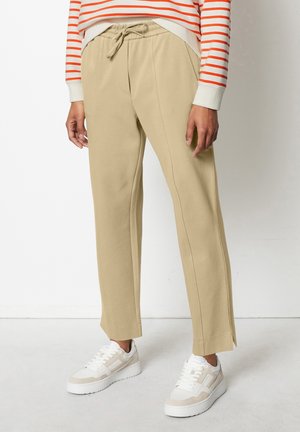 Persona con pantalones beige con cordón y zapatillas blancas de pie, con parte de una camiseta de manga larga a rayas rojas y blancas visible.