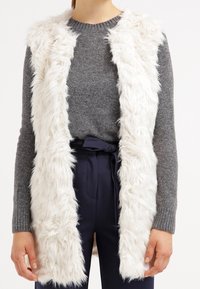 Faux fur utan ärm väst i vitt, med en plysch-liknande textur, öppen framsida och mjukt foder. Buren över en grå tröja och marinblå byxor.