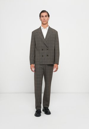 Man standing against white background wearing brown checked double-breasted suit, white shirt, and black shoes.