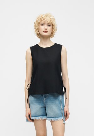 Göndör szőke hajú nő, aki ujjatlan fekete felsőt visel oldalt megkötve és rojtos farmer rövidnadrágot, egy egyszínű fehér háttér előtt áll.