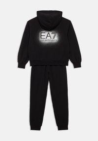 Črni športni trenirka, ki vključuje kapucar s kapuco in ožje krojene hlače. Na hrbtni strani ima srebrn logotip. Izdelan je iz mehke tkanine.