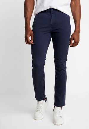 Pantalons slim-fit bleu marine en tissu léger, dotés de cinq poches, d'passants de ceinture et d'une texture lisse, accompagnés de baskets blanches.