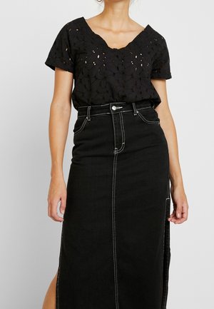 Femme portant un chemisier noir à manches courtes en dentelle florale, rentré dans une jupe en jean noire taille haute avec couture blanche et fente latérale.