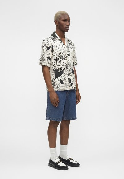 Άνδρας με κοντά ξανθά μαλλιά φορώντας ένα πουκάμισο με σχέδια και κοντά μανίκια, τζιν σορτς μέχρι το γόνατο, λευκές κάλτσες και ασπρόμαυρα μοκασίνια.