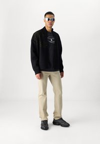 Sudadera negra con cremallera y líneas amarillas, camiseta negra con logo, pantalones beiges y zapatillas negras. Unas gafas de sol reflectantes completan el look.