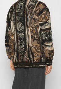 Sweter dziany z abstrakcyjnymi wzorami w kolorach brązowym, czarnym i beżowym. Charakteryzuje się luźnym krojem, prążkowanymi mankietami i dołem oraz teksturowanym wzorem.