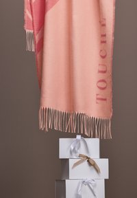 Écharpe couleur pêche avec bords frangés, présentant un motif à rayures diagonales et le mot "TOUCHÉ" dans une teinte plus foncée, drapée sur des boîtes cadeaux.