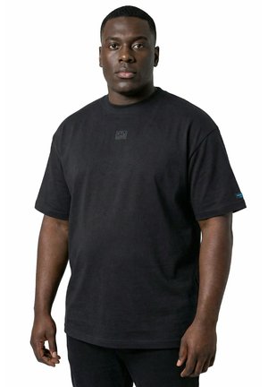 Hombre negro que lleva una camiseta negra sencilla de manga corta con un pequeño logo en el pecho y una etiqueta azul en la manga.