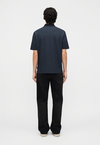 Άνδρας με τα πίσω γυρισμένα, φοράει κοντομάνικο πουκάμισο σκούρου μπλε με σχέδιο, μαύρα τζιν και ανοιχτόχρωμα παπούτσια σε απλό φόντο.