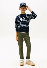 Tamsiai mėlynas dryžuotas džemperis su baltomis raidėmis, alyvuogių spalvos kelnės ir smėlio spalvos sportbačiai. Kepurė tamsiai mėlyna su baltais užrašais.