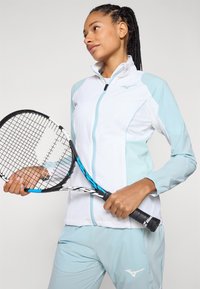Leichte Tennisjacke in Weiß und Hellblau, mit Reißverschluss, Stehkragen und elastischen Bündchen, kombiniert mit passenden Hosen.